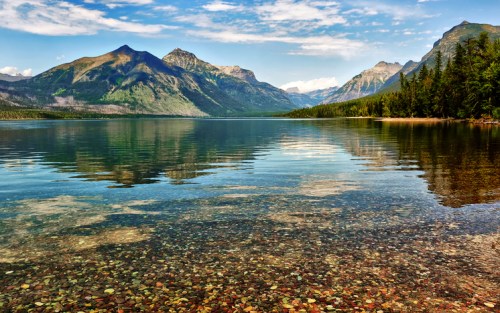 A serene lake reflects mountains and a clear sky, with colorful stones visible beneath the calm water's surface.