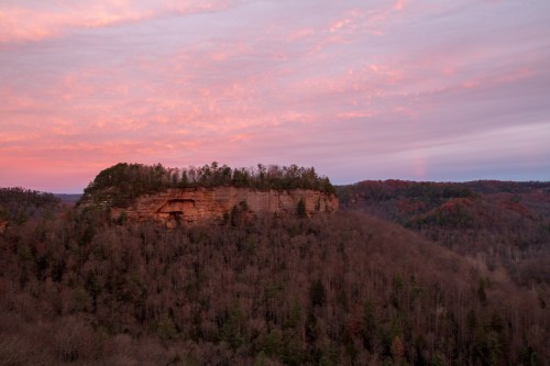 A scenic view of a rocky cliff surrounded by trees, with a colorful sunset sky in the background.