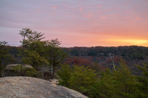 A scenic view of a rocky landscape at sunset, with trees and colorful clouds in the sky.