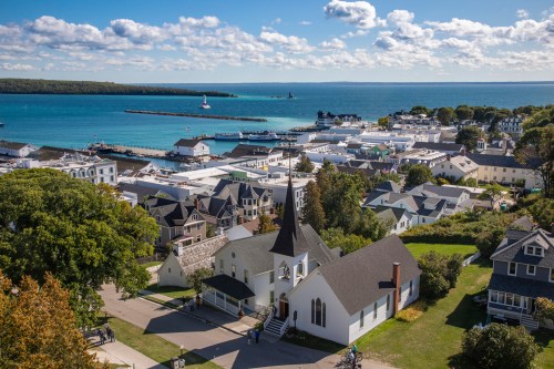 A scenic view of a coastal town with a church steeple, harbor, and boats under a blue sky with fluffy clouds.