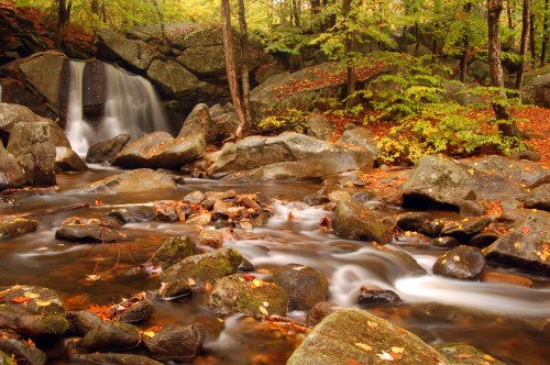 A serene waterfall cascades over rocks, surrounded by autumn foliage and a gently flowing stream.