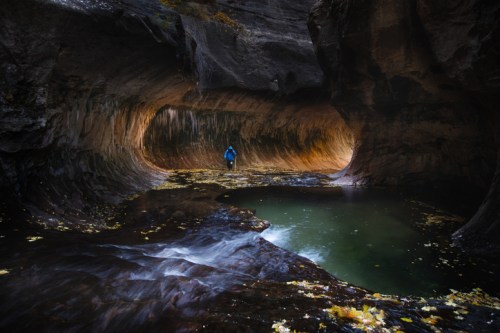 A person stands in a narrow, rocky canyon with flowing water and autumn leaves scattered on the ground.