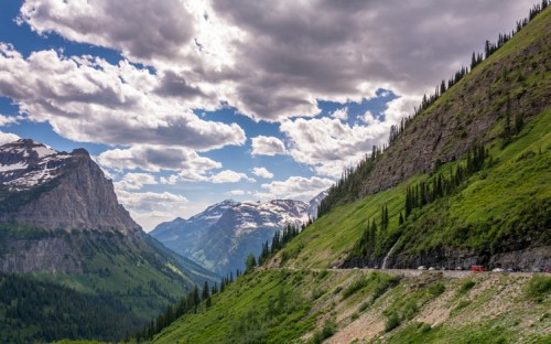 A scenic mountain landscape with lush greenery, snow-capped peaks, and a winding road under a cloudy sky.