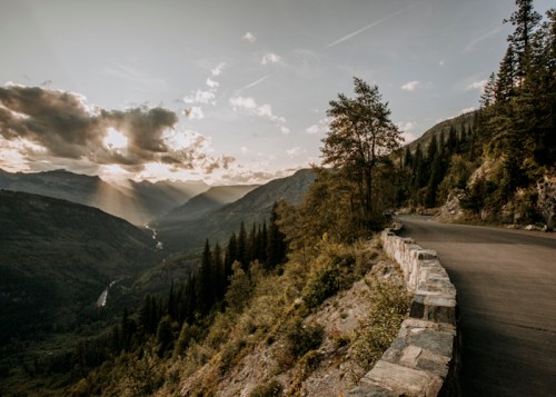 A scenic mountain view with a winding road, lush trees, and sunlight breaking through clouds over a valley.