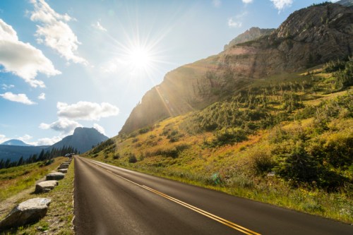 A sunlit road winds through a mountainous landscape, surrounded by greenery and rocky cliffs under a blue sky.