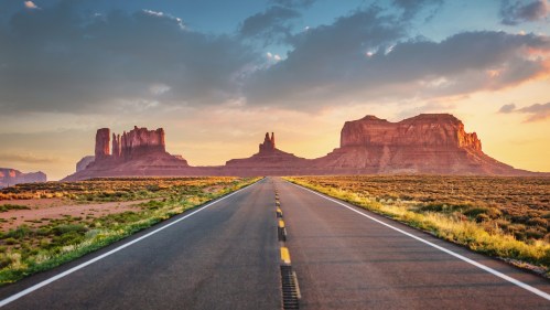 A scenic road leads through a desert landscape with towering rock formations under a colorful sky at sunset.