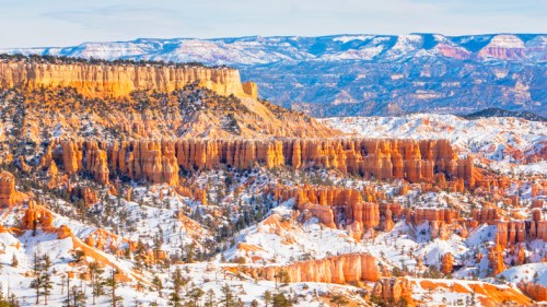 A snowy landscape of Bryce Canyon, featuring orange rock formations and distant mountains under a clear sky.