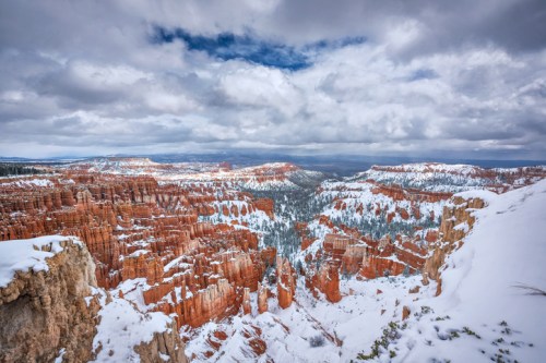 A snowy landscape of Bryce Canyon, showcasing red rock formations under a cloudy sky.