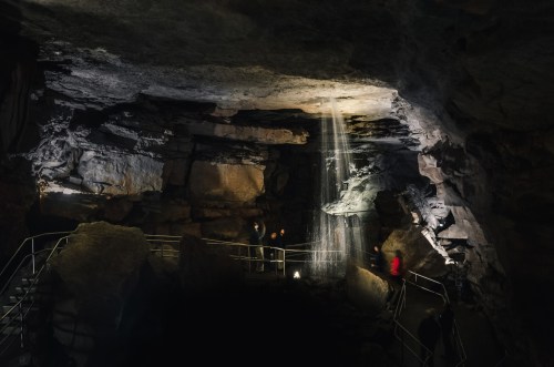 A dimly lit cave with rock formations and a beam of light illuminating a waterfall, with visitors exploring the area.