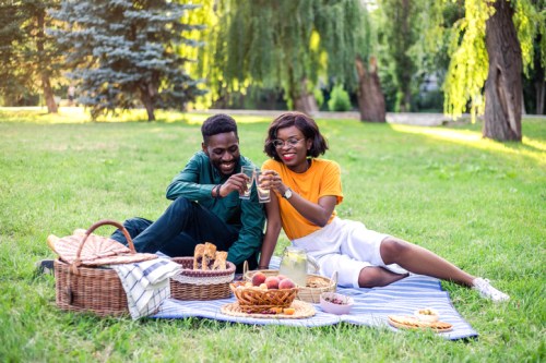 A couple enjoys a picnic on a grassy field, smiling and sharing drinks with food spread around them.