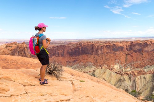 A woman in a pink cap and backpack stands on a rocky ledge, overlooking a vast canyon landscape under a blue sky.