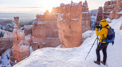 A photographer in a yellow jacket captures a snowy landscape at sunrise in a canyon with towering rock formations.