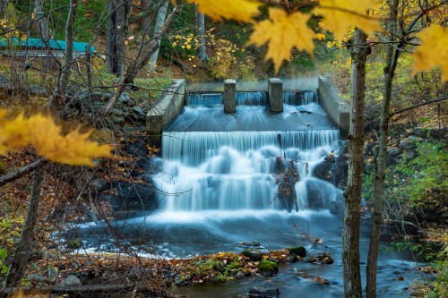 A serene waterfall cascades over a dam, surrounded by autumn foliage and rocky terrain.