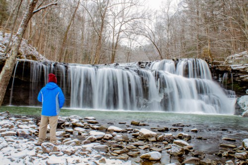 A person in a blue jacket and red hat stands by a snowy waterfall, surrounded by trees and rocky terrain.