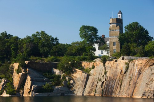A lighthouse stands atop rocky cliffs, surrounded by greenery and a clear blue sky, overlooking calm waters.