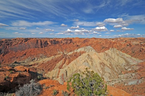 Vast desert landscape with layered red and orange rock formations under a blue sky with fluffy white clouds.