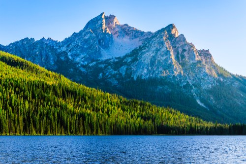 A serene mountain landscape with a lake in the foreground and dense evergreen trees lining the shore.