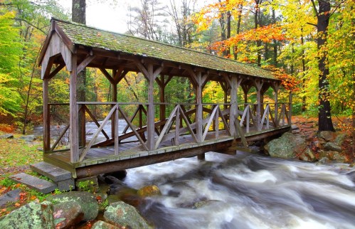 A wooden covered bridge spans a rushing stream, surrounded by vibrant autumn foliage and rocky terrain.