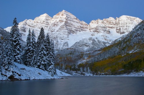 Snow-capped mountains rise above a serene lake, surrounded by evergreen trees and autumn-colored foliage.