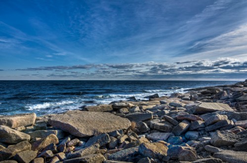Rocky shoreline with waves lapping against the stones under a blue sky with scattered clouds.