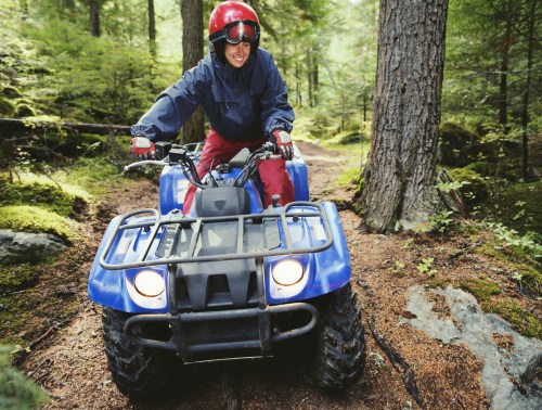 A person riding a blue ATV on a forest trail, wearing a red helmet and smiling amidst lush greenery.