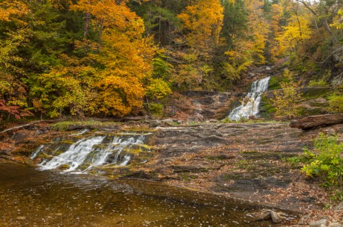 A serene waterfall cascades over rocky terrain, surrounded by vibrant autumn foliage in shades of orange and yellow.