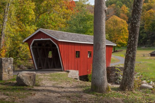 A red covered bridge surrounded by trees with autumn foliage, set in a peaceful natural landscape.