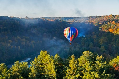 A colorful hot air balloon floats over a lush, green valley with trees and a river below, surrounded by autumn foliage.