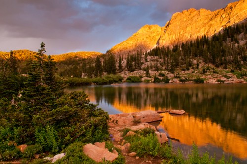A serene lake reflects golden mountain peaks and lush greenery under a dramatic sky at sunset.