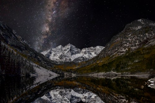 A starry night sky reflects over a serene lake, surrounded by snow-capped mountains and autumn-colored trees.
