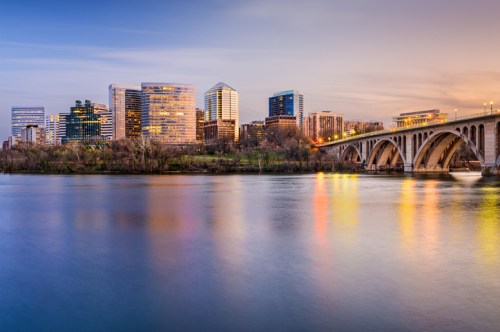 City skyline at dusk with modern buildings and a bridge over a calm river, reflecting colorful lights.