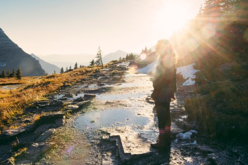 A person stands on a rocky path at sunset, surrounded by mountains and trees, with sunlight creating a lens flare effect.