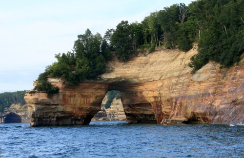 Natural rock formation with an arch, surrounded by water and lush greenery, showcasing layered geological colors.