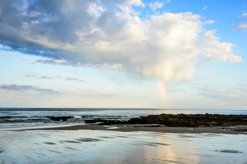 A serene beach scene with gentle waves, rocky shore, and a rainbow emerging from clouds in a blue sky.