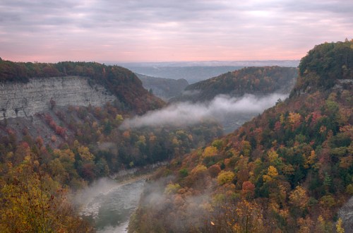 A misty valley surrounded by colorful autumn foliage and steep cliffs under a cloudy sky.