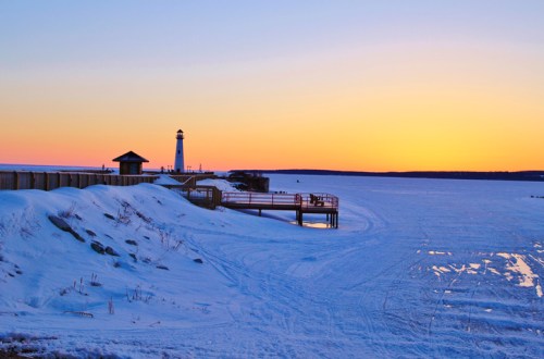 A snowy shoreline at sunset, featuring a lighthouse and calm waters reflecting the colorful sky.