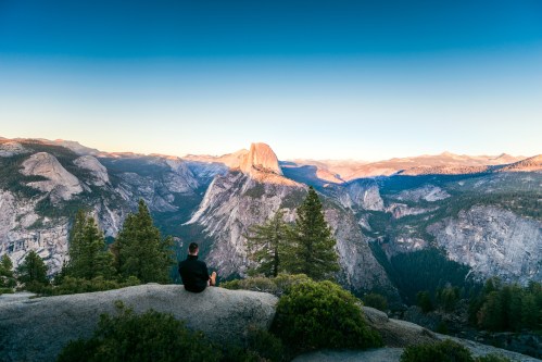 A person sits on a rock, gazing at a vast mountain landscape during sunset, with trees in the foreground.
