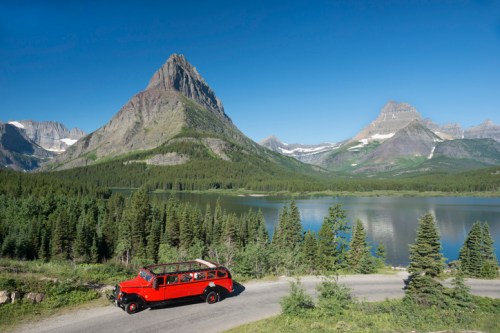 A vintage red bus parked by a serene lake, surrounded by lush forests and majestic mountains under a clear blue sky.