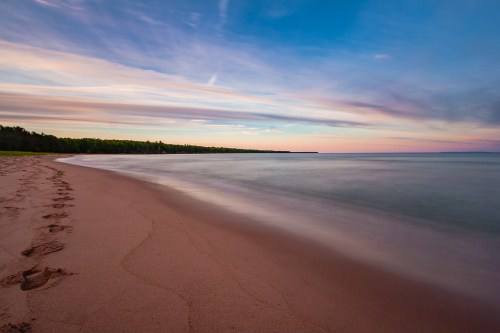 A serene beach at sunset, with gentle waves and footprints in the sand under a colorful sky.