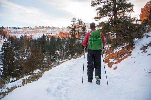 A hiker with a green backpack walks on a snowy trail surrounded by trees and rocky cliffs.