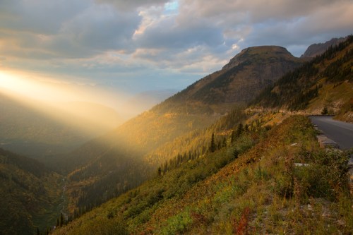 Sunlight streams through clouds over a mountainous landscape, highlighting autumn foliage along a winding road.