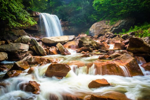 A serene waterfall cascades over rocks, surrounded by lush greenery and flowing water in a tranquil natural setting.
