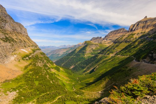 A breathtaking view of a lush green valley surrounded by towering mountains under a blue sky with wispy clouds.