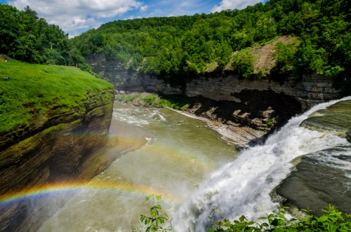 A vibrant waterfall cascades into a river, surrounded by lush green cliffs and a rainbow arching over the scene.
