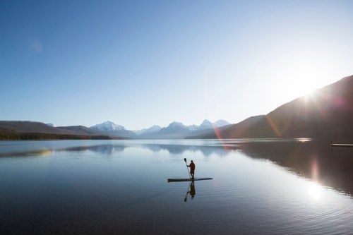 A person stands on a paddleboard in a calm lake, surrounded by mountains under a clear blue sky.