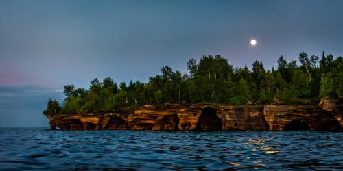 A serene lakeside view at dusk, featuring rocky cliffs, lush trees, and a full moon illuminating the sky.