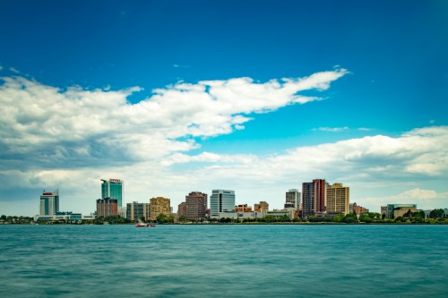 A skyline view of a city across a river, featuring modern buildings under a blue sky with scattered clouds.