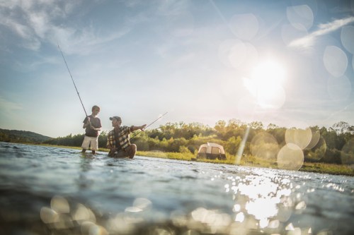 A man and a child fishing in a river under a sunny sky, with a camper in the background.