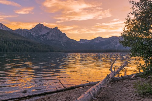 A serene lake at sunset, reflecting vibrant colors with mountains in the background and a log on the shore.