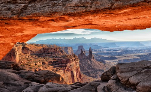 A stunning view of rugged cliffs and distant mountains framed by a rock overhang in a desert landscape.